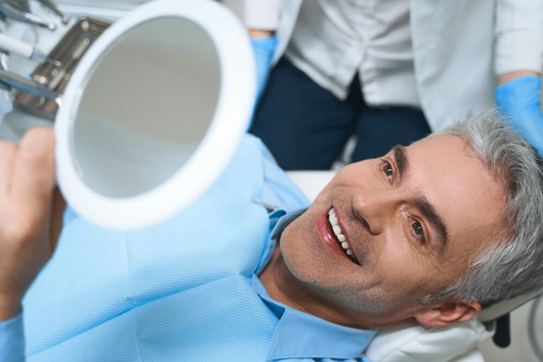 A male dental patient lays back in a dental exam chair and admires his smile in a handheld mirror